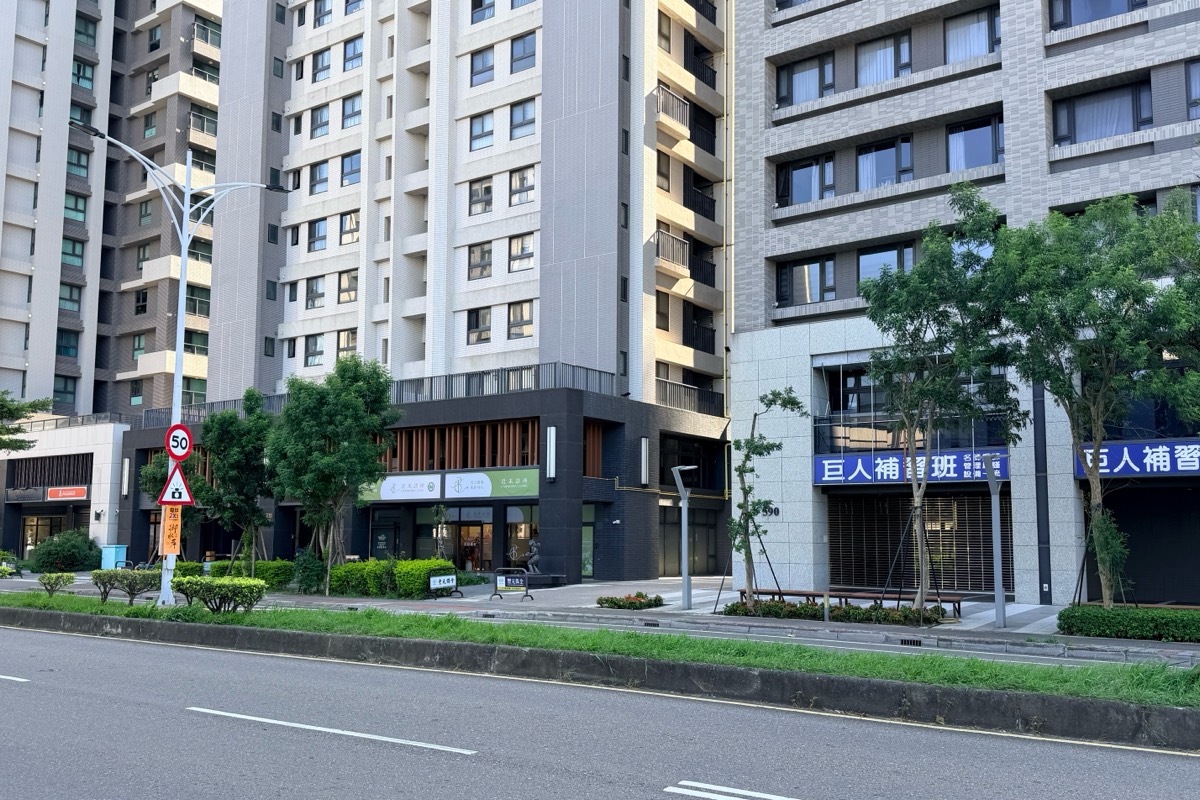 從中山路一段對面望向診所所在的大樓：幾棟高樓並排，其中一棟一樓是深色外牆、上方有木色直條格柵、掛著淺綠色招牌的店面，右側大樓一樓為巨人補習班。
