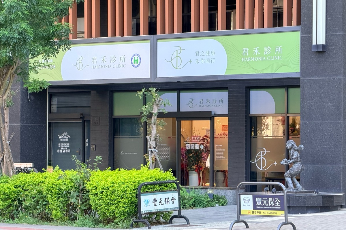 從對街看君禾診所門面：二樓有木色直條格柵，一樓上方是淺綠色的君禾診所招牌與「君之健康 禾你同行」字樣，落地玻璃門內擺著花籃，右側有一座小女孩銅像，人行道上有保全公司的禁止停車立牌。