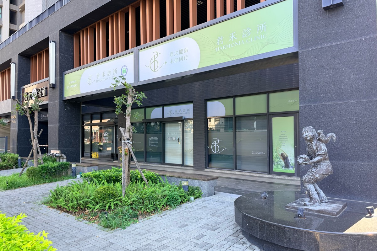 站在人行道上看君禾診所：招牌寫著「君禾診所 HARMONIA CLINIC」與「君之健康 禾你同行」，騎樓下是整排落地玻璃窗，前方有植栽，右側是坐在石台上的小女孩銅像。