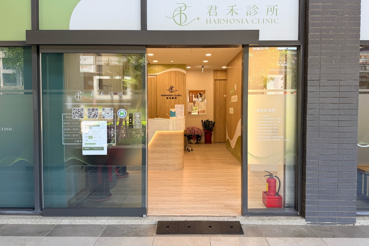君禾診所門面：上方是淺綠色的君禾診所招牌，中間的自動玻璃門敞開，門口地上橫鋪著一塊黑色斜坡板，門內可見櫃檯與木質牆面，左側玻璃上貼著門診時間表與 QR 碼。